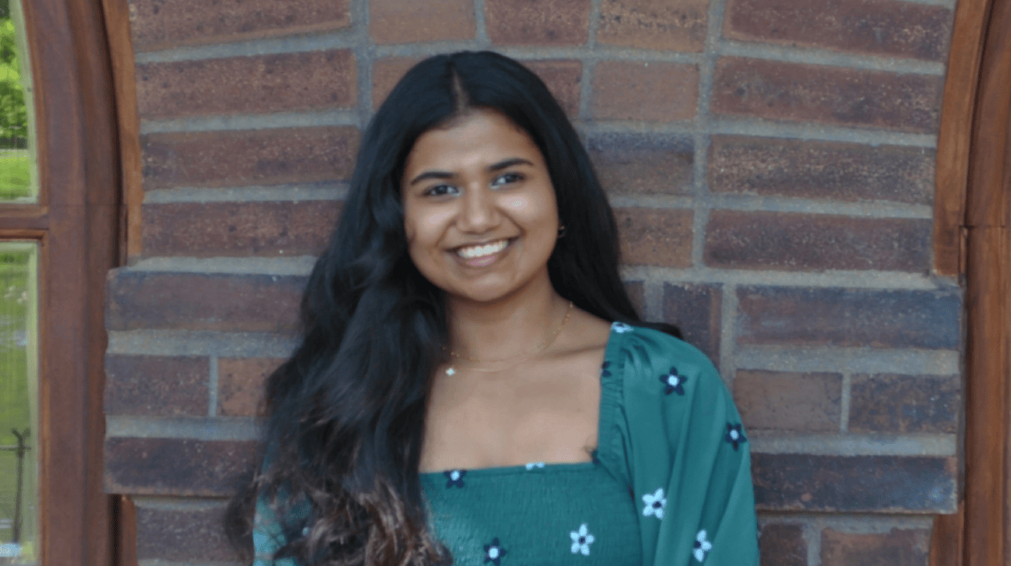 Young woman smiling in front of a brick wall, with long dark hair and wearing a green floral top with a square neckline.